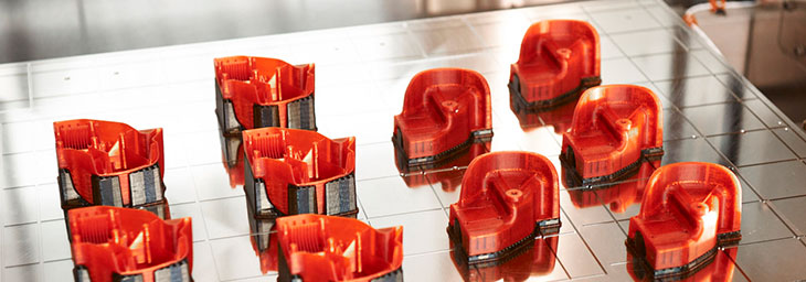 A imagem mostra 10 peças de coloração vermelha e detalhes em metal, impressas em 3D. Elas estão expostas sob uma bancada de metal.