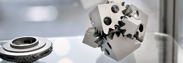 A imagem mostra dois objetos impressos em metal. Uma das peças é redonda com encaixes na parte superior e outra tem formato de cubo, com alguns furos na sua estrutura.