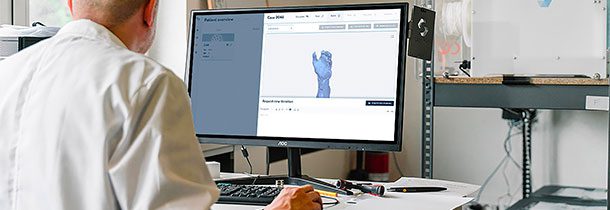 A imagem mostra um homem caucasiano que está de costas sentado e em frente a uma mesa com computador. Na tela está aberto um software com um desenho digital. Ele veste jaleco branco. Ao fundo, está uma impressora 3D.