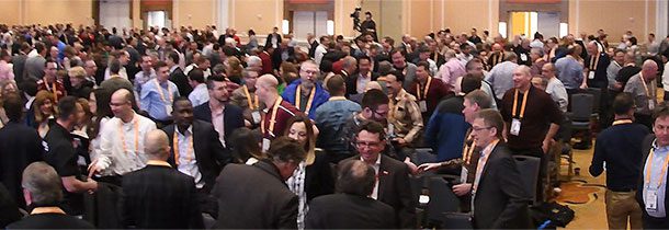 A imagem mostra diversas pessoas na AMUG Conference. A multidão está de pé em uma sala com paredes amarelas.