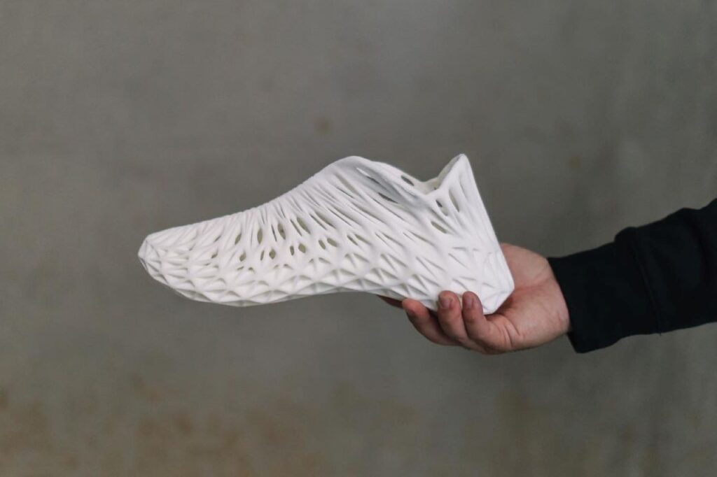 A imagem mostra uma mão segurando um tênis totalmente branco, com design vazado, feito por impressão 3D. O fundo da imagem é cinza. 