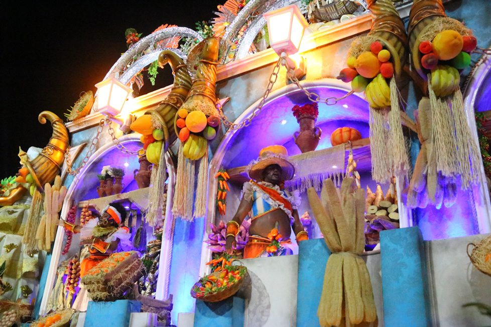 Impressora 3D industrial produzindo peça complexa em ambiente de engenharia, com projetos digitais exibidos em monitores ao fundo